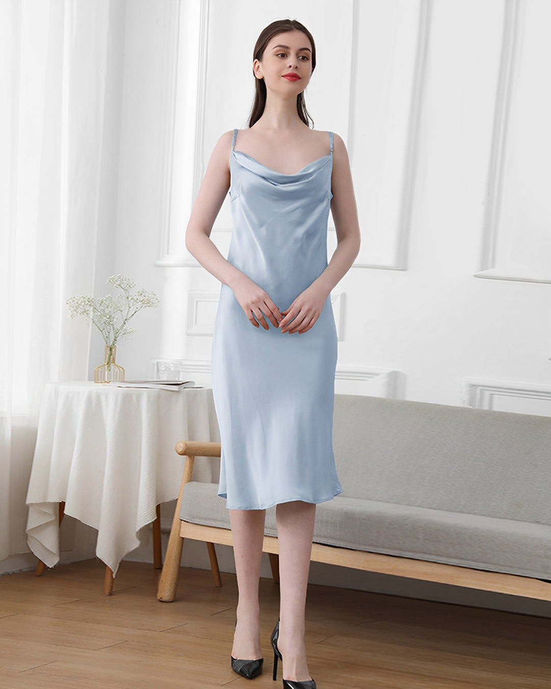 A woman wearing the Elegant Cowl Neck Silk Nightdress from SusanSilk stands in a minimalist room with white walls, a small table, and a bench.