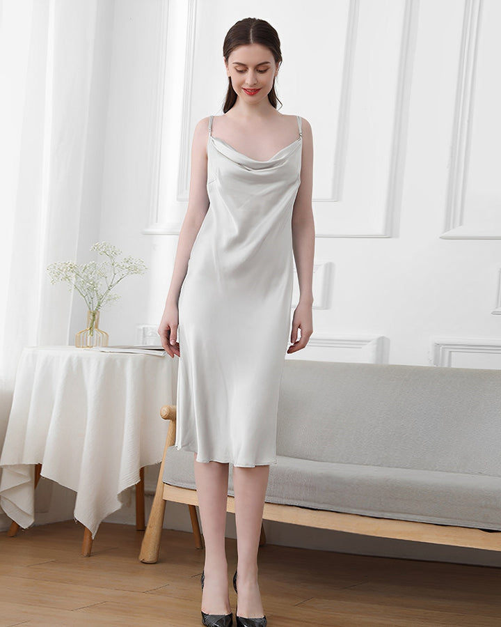 A woman stands indoors wearing an Elegant Cowl Neck Silk Nightdress by SusanSilk, next to a small round table with a white tablecloth. She is smiling slightly, with her hands by her sides.