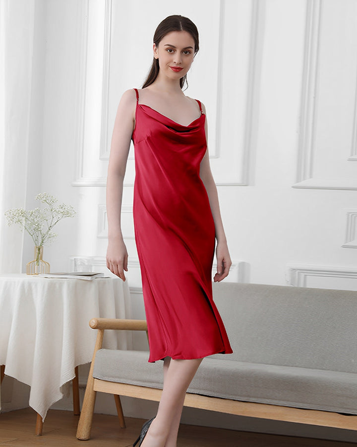 A woman in an Elegant Cowl Neck Silk Nightdress by SusanSilk stands indoors near a gray sofa and a small table with flowers.