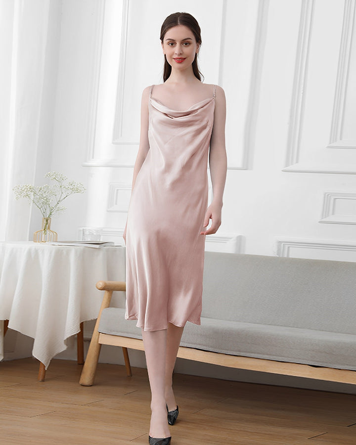 A woman dressed in an Elegant Cowl Neck Silk Nightdress by SusanSilk stands in a white room with a table and a couch.