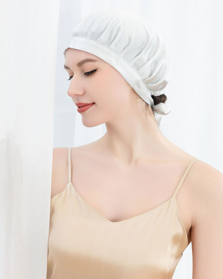 A woman wearing a Chic Silk Bonnet For Sleeping with Ribbons and a beige camisole from SusanSilk looks down while standing against a white curtain.