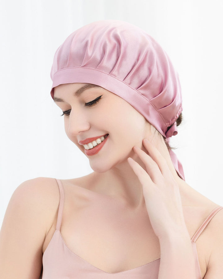 A person wearing a SusanSilk Chic Silk Bonnet For Sleeping with Ribbons and a matching dress smiles while touching their neck in a softly lit setting, basking in the luxurious feel of silk.