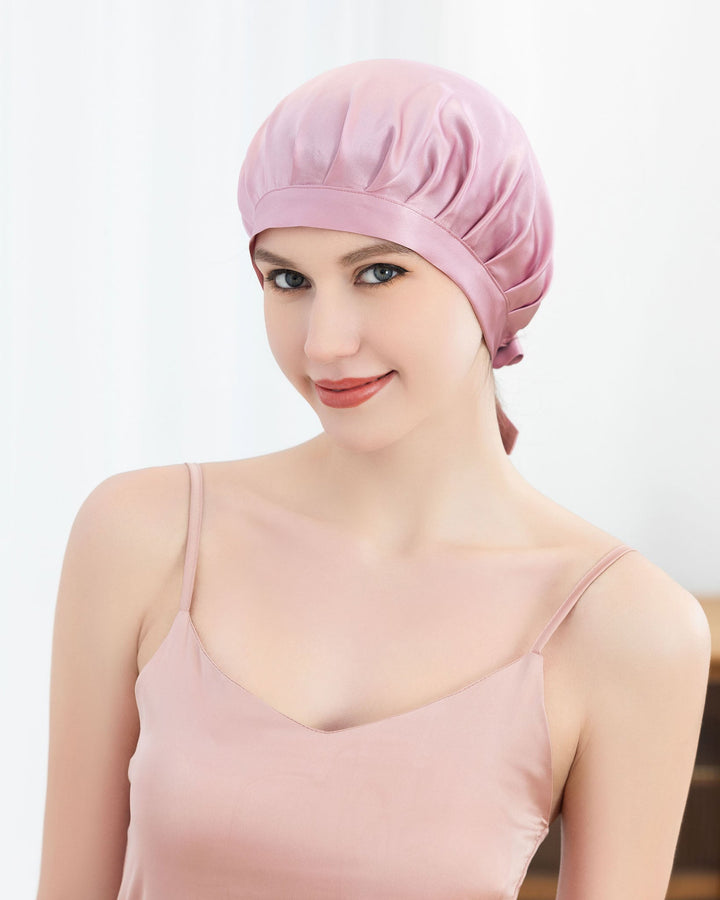 A woman wearing a pink Chic Silk Bonnet For Sleeping with Ribbons and a SusanSilk sleeveless top poses and smiles against a plain background.