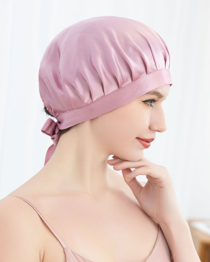 A person is shown in profile, touching their chin with one hand while wearing a pink Chic Silk Bonnet For Sleeping with Ribbons from SusanSilk and a pink camisole.