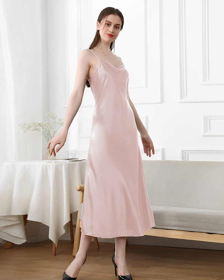 A person wearing the SusanSilk 19 Momme Silk Sling Dress For Women in light pink strikes a pose in a room with white walls, a table adorned with a white tablecloth, and a light-colored couch.