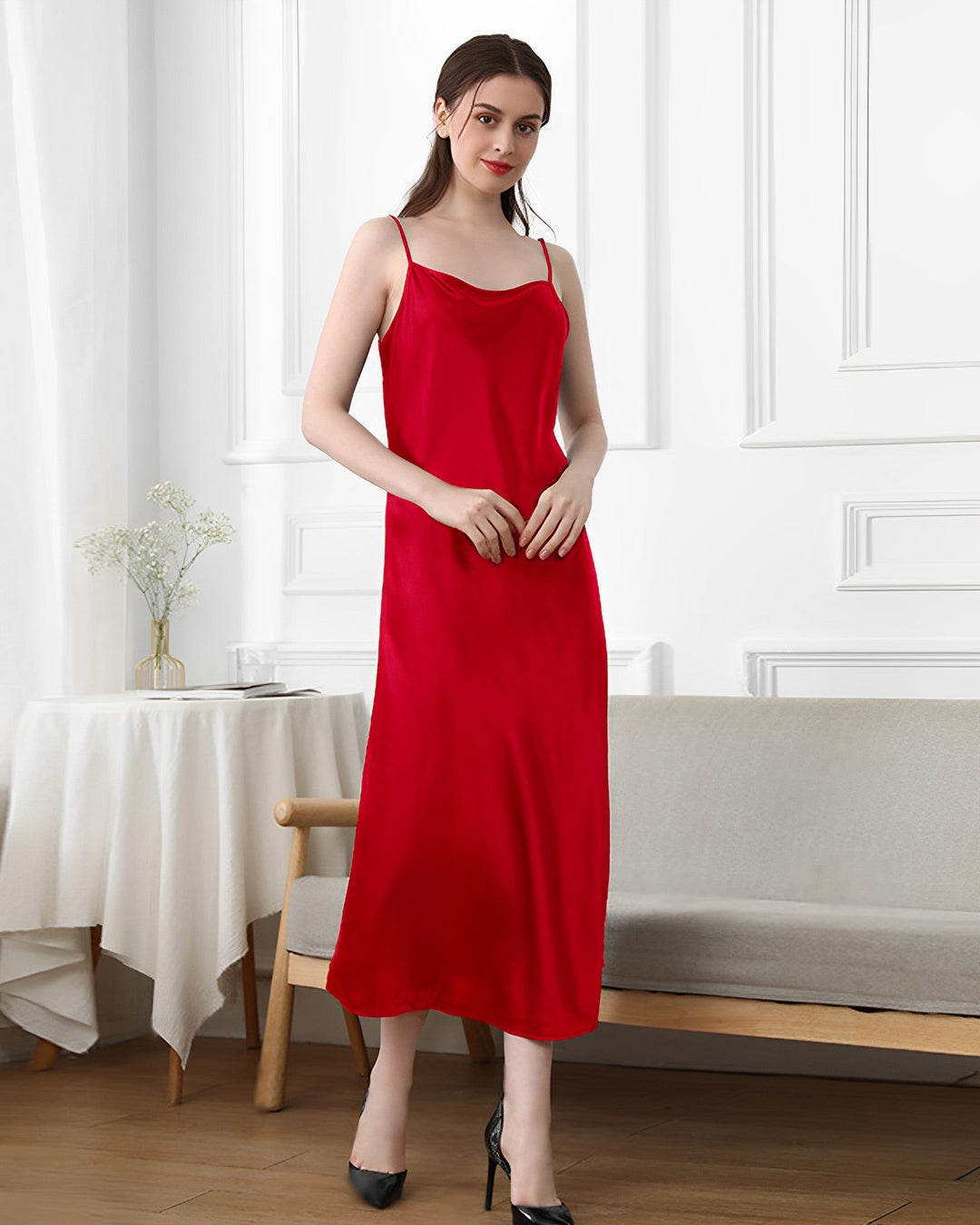 A woman in a red 19 Momme Silk Sling Dress for women from SusanSilk stands indoors next to a grey bench and a table with a white cloth and susansilk flowers.