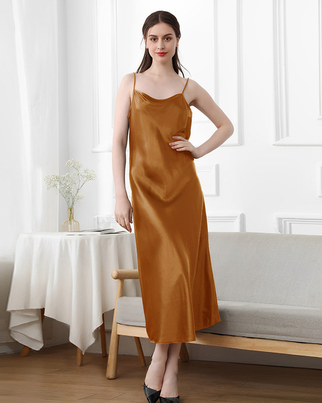 A woman wearing the 19 Momme Silk Sling Dress for Women by SusanSilk stands in a minimalistic room with white walls, a light grey couch, and a small table with a white cloth and vase.