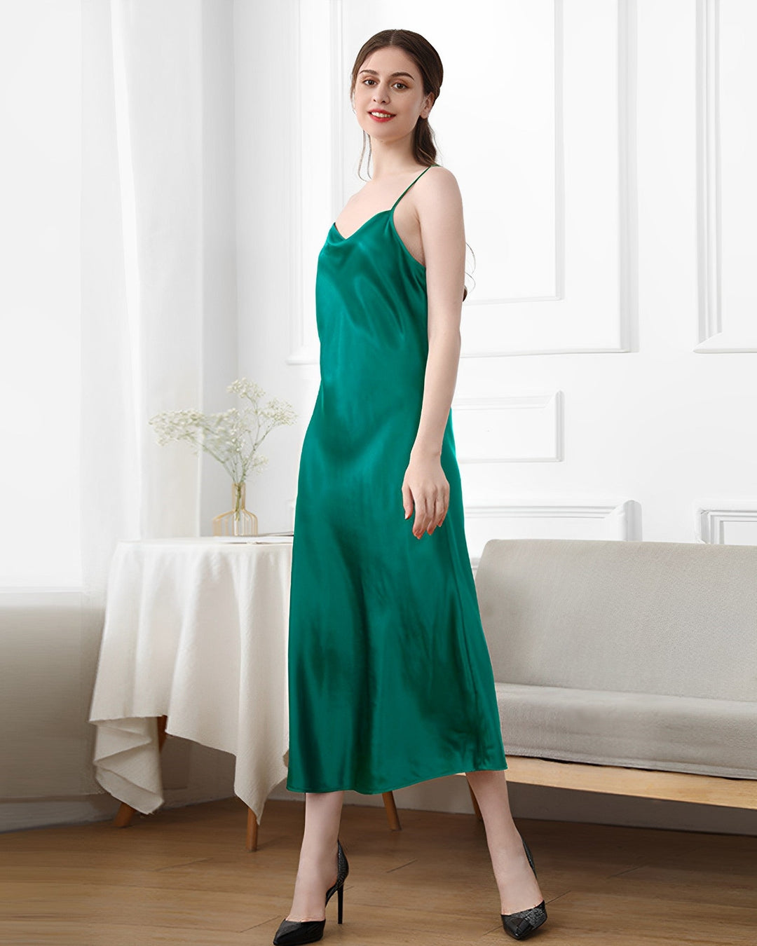 A woman stands in a bright room, wearing the 19 Momme Silk Sling Dress for Women by SusanSilk in teal, paired with black high heels. She poses gracefully, with one hand resting on a bench and the other by her side. A table adorned with a white cloth and a plant can be seen in the background, accentuating the timeless elegance of her SusanSilk attire.