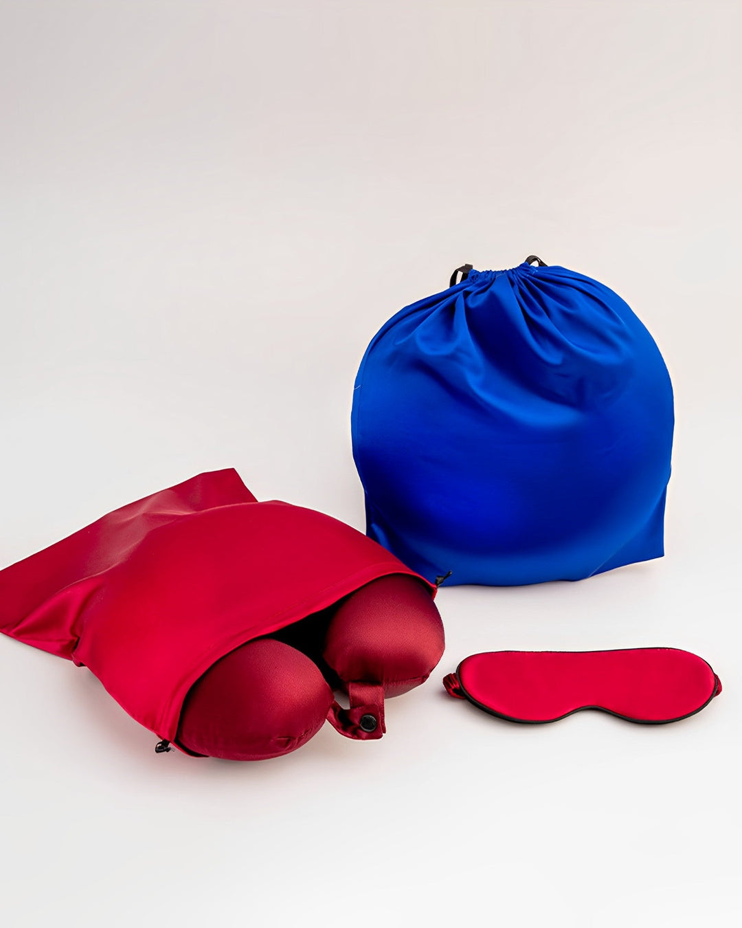 A red bag containing two spherical objects, a blue drawstring bag, and a red silk eye mask from the SusanSilk 19 Momme Silk U-Shaped Pillow Travel Set are placed on a light background.