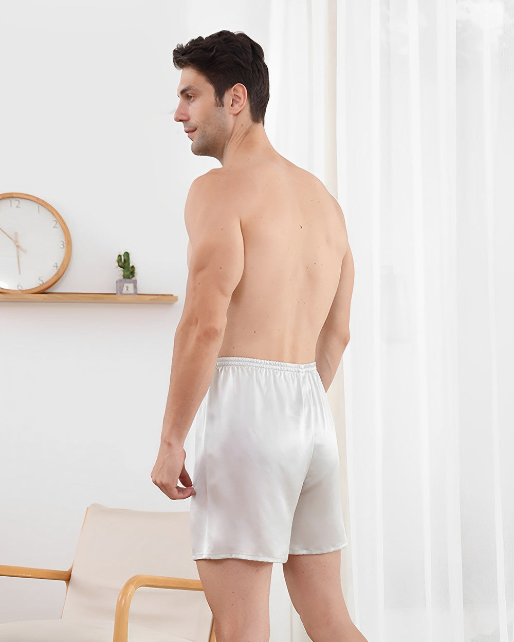 A shirtless man stands in a room wearing SusanSilk's 19 Momme Comfortable Silk Boxer, facing left. A wall clock and a small potted plant sit on a shelf behind him, next to a beige armchair.