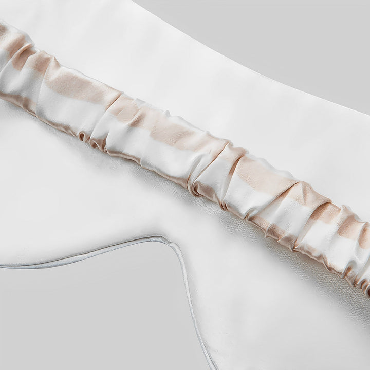 Close-up of the 19 Momme Stripe Silk Sleep Eye Mask by SusanSilk, showcasing its white satin fabric with ruched elastic detail and beige accents on a plain background.