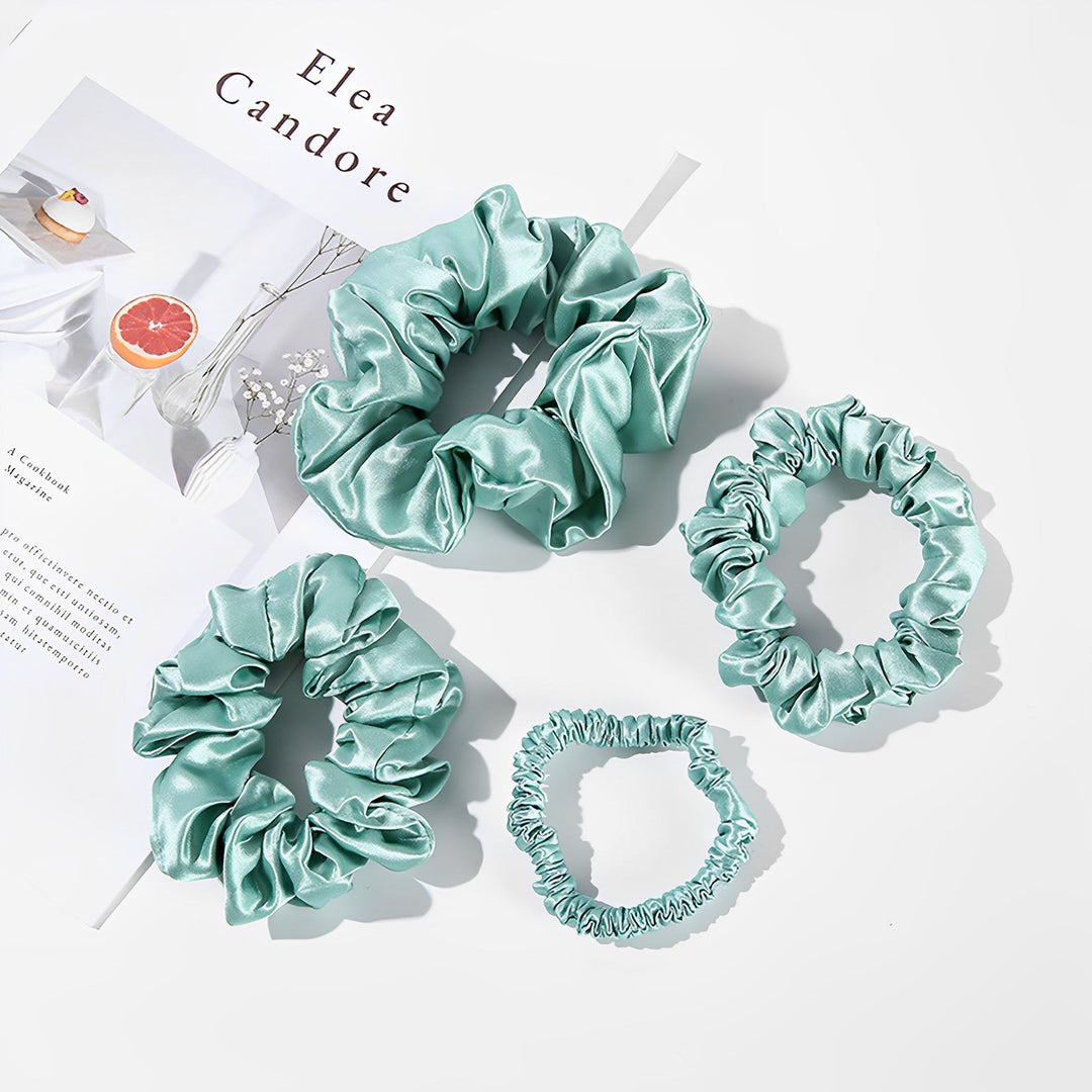 Four 22 Momme Luxury Silk Scrunchies from SusanSilk, in light blue and varying sizes, are displayed on a white background next to a magazine page featuring a grapefruit and a plant.