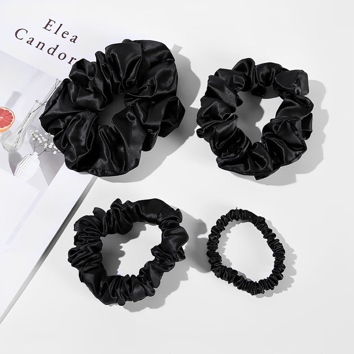 Four black silk scrunchies of different sizes are laid out on a white background next to a magazine, showcasing the luxury and elegance of the 22 Momme Luxury Silk Scrunchies Set 4-Pack by SusanSilk.