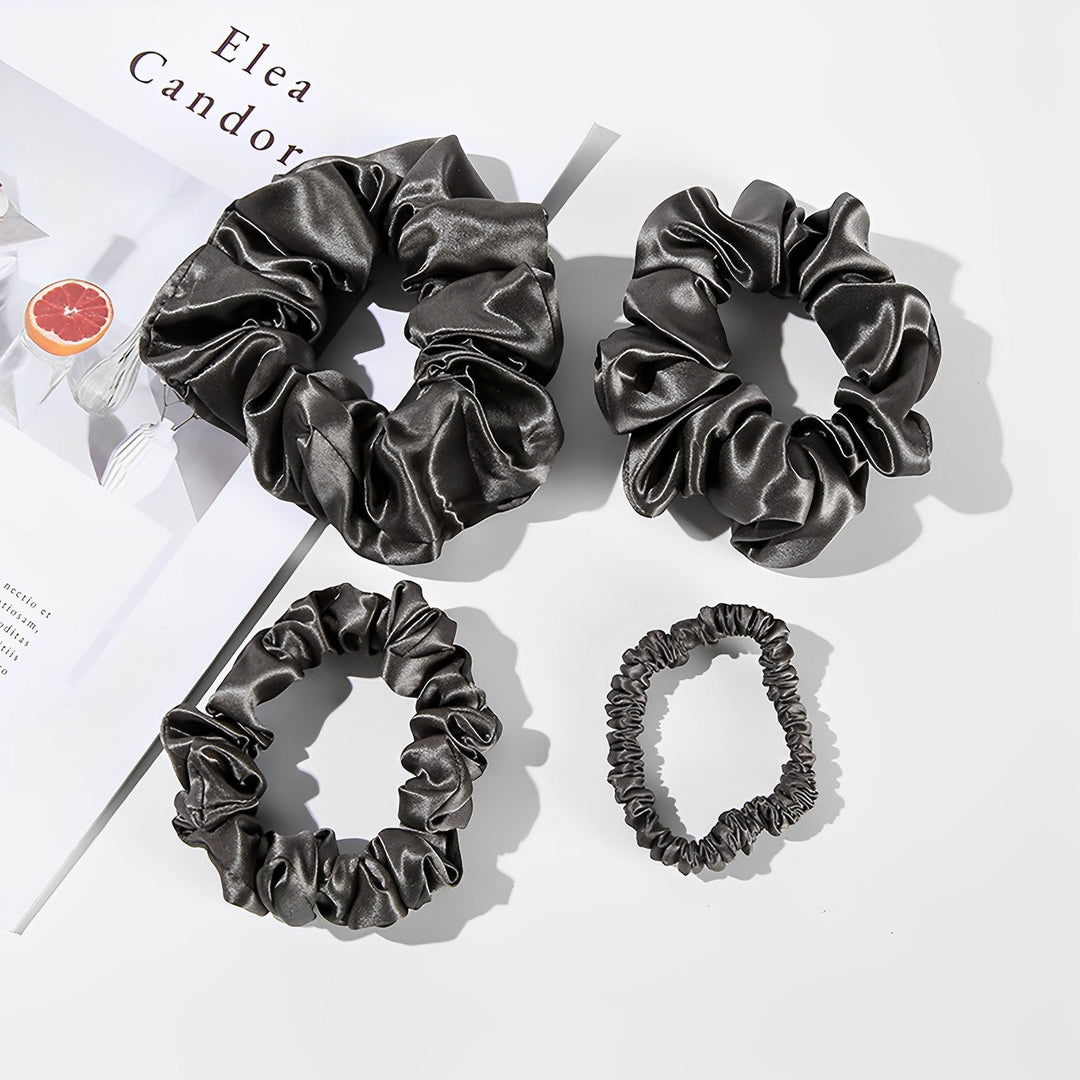 Four luxurious black satin scrunchies of varying sizes are artfully arranged on a flat white surface next to a magazine with a grapefruit image and the text "Elea Candor." The 22 Momme Luxury Silk Scrunchies Set 4-Pack by SusanSilk adds an elegant touch to your hair accessory collection, reminiscent of their renowned quality.