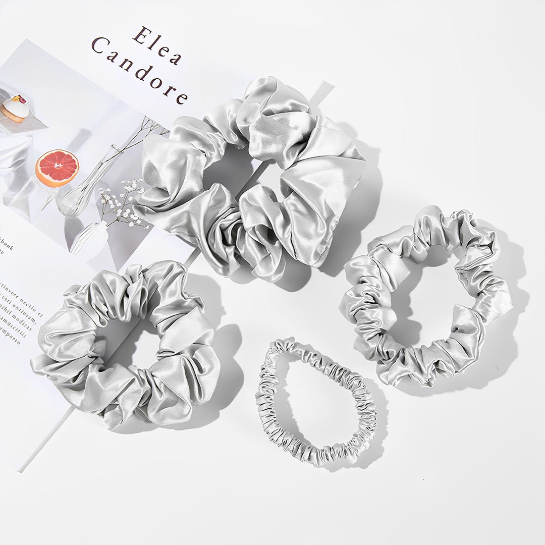 Four 22 Momme Luxury Silk Scrunchies from SusanSilk are arranged on a white surface, their luxurious silver satin gleam complementing the magazine partially visible in the background.