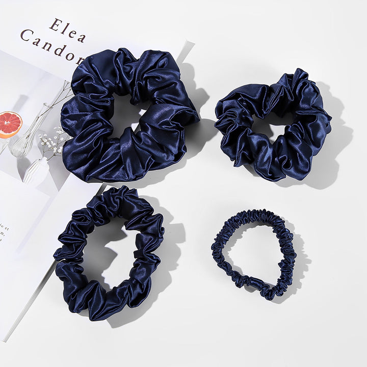 Four 22 Momme Luxury Silk Scrunchies from SusanSilk, all in navy blue and of varying sizes, are displayed on a white surface next to an open magazine.