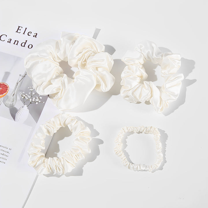 The 22 Momme Luxury Silk Scrunchies Set 4-Pack from SusanSilk features varying sizes of white satin scrunchies displayed on a white surface beside a minimalistic SusanSilk magazine.