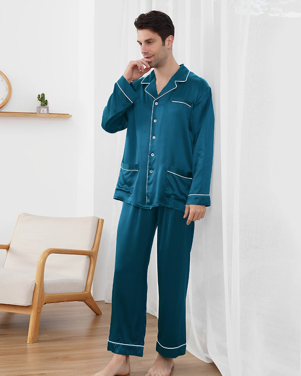 A man is standing in a room wearing SusanSilk's 22 Momme Lapel Collar Silk Pajamas Set in teal with white piping, the luxurious fabric reminiscent of susansilk. He is looking to the side and touching his lips. There is a chair, shelf, and small clock in the background.