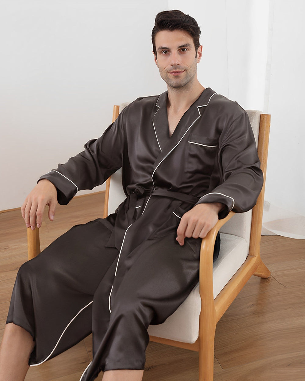 A man sitting in a chair wearing a SusanSilk 22 Momme Luxury Silk Robe with white piping details, exuding an air of refinement.