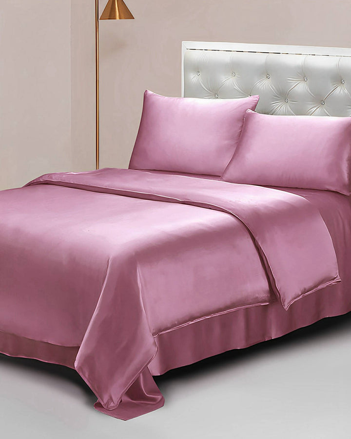 A neatly made bed features SusanSilk's 22 Momme Seamless Silk Duvet Cover with matching pillowcases and a coverlet, all in a luxurious pink satin. The headboard is light-colored with tufted upholstery, and a tall, gold lamp stands gracefully on the left side, adding an extra touch of elegance to the serene setup.