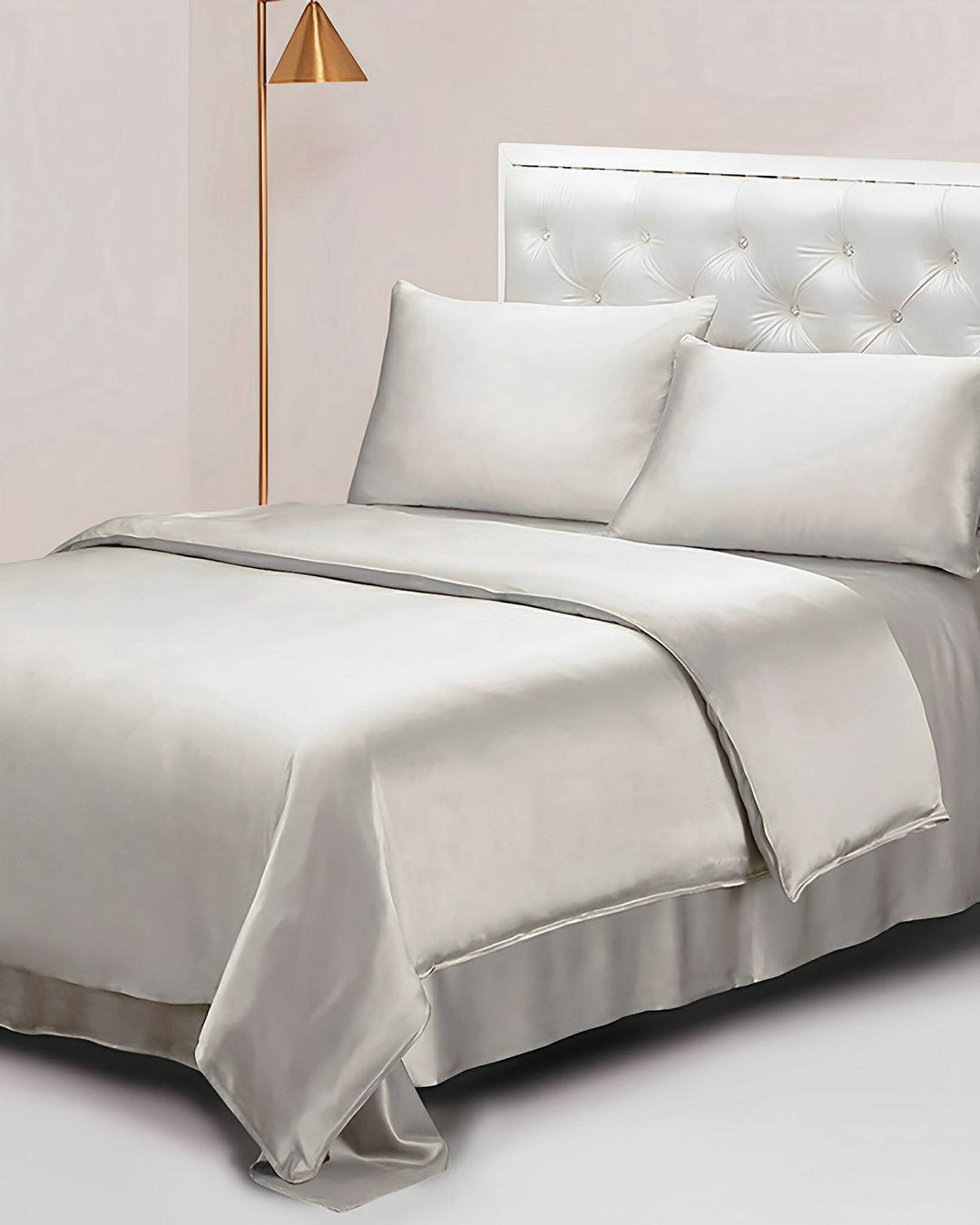 A neatly made bed featuring the 25 Momme Seamless Luxury Duvet Cover by SusanSilk, crafted from smooth silk in light gray, including two pillows, a quilt, and a comforter, is flanked by a gold floor lamp and a white tufted headboard.