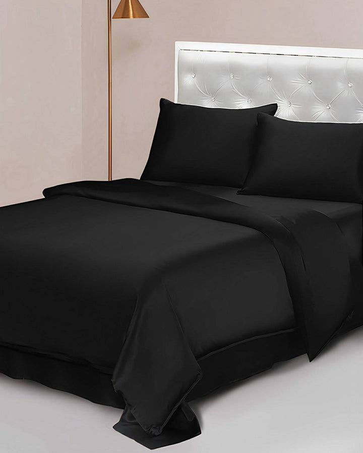 A neatly made bed featuring a 25 Momme Seamless Luxury Duvet Cover by SusanSilk, accompanied by two matching pillows, a black comforter, and a white tufted headboard. Beside the bed stands a floor lamp.