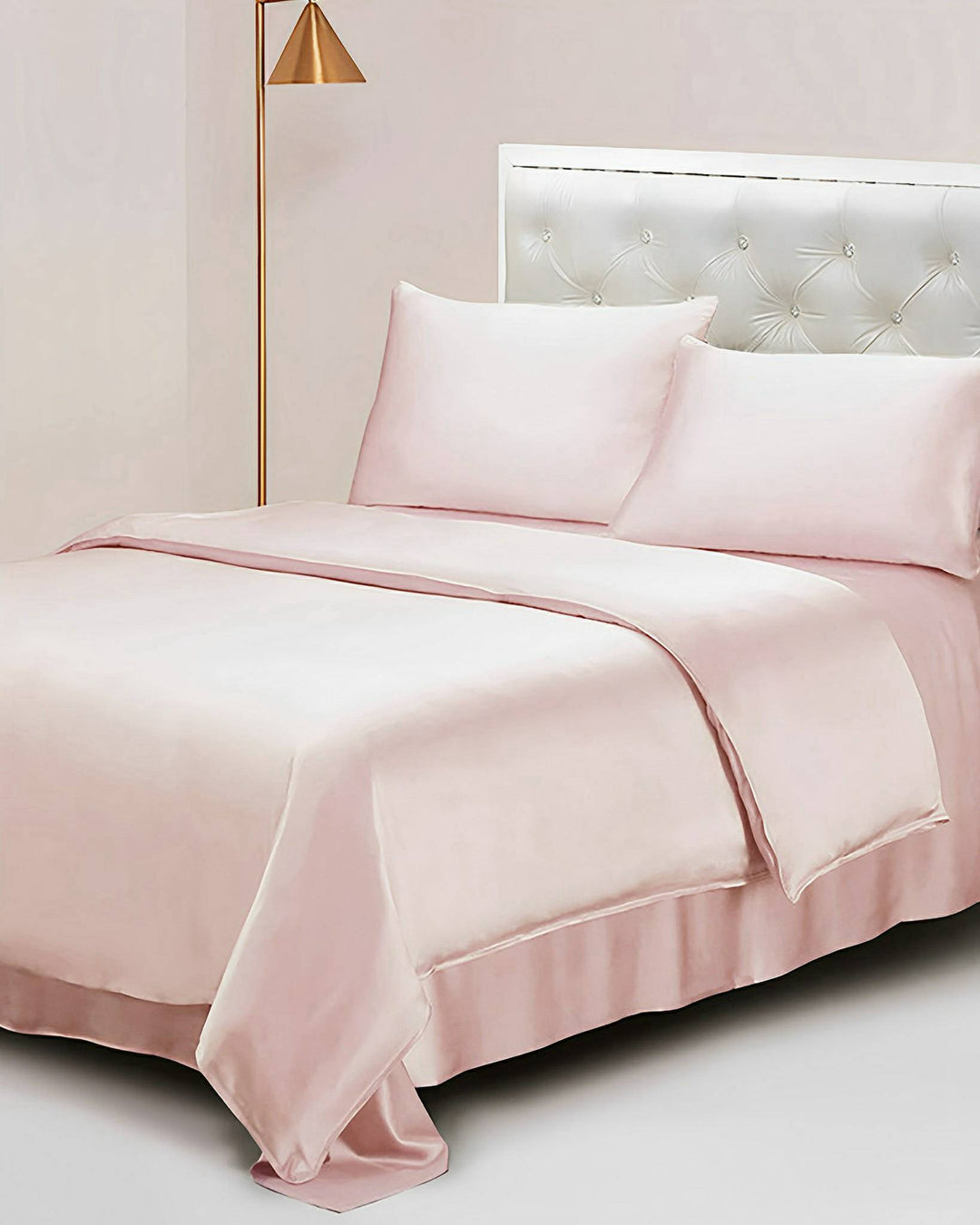 A neatly made bed with the 25 Momme Seamless Luxury Duvet Cover by SusanSilk in a pale pink shade, complemented by matching pillowcases, a tufted white headboard, and a gold floor lamp on the side.