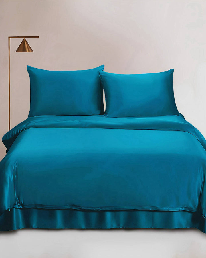 A neatly made bed showcasing the SusanSilk 25 Momme Seamless Luxury Duvet Cover in a rich teal silk, complemented by two pillows, a comforter, and a sheet, is displayed. To the left side of the bed stands a modern brass floor lamp.