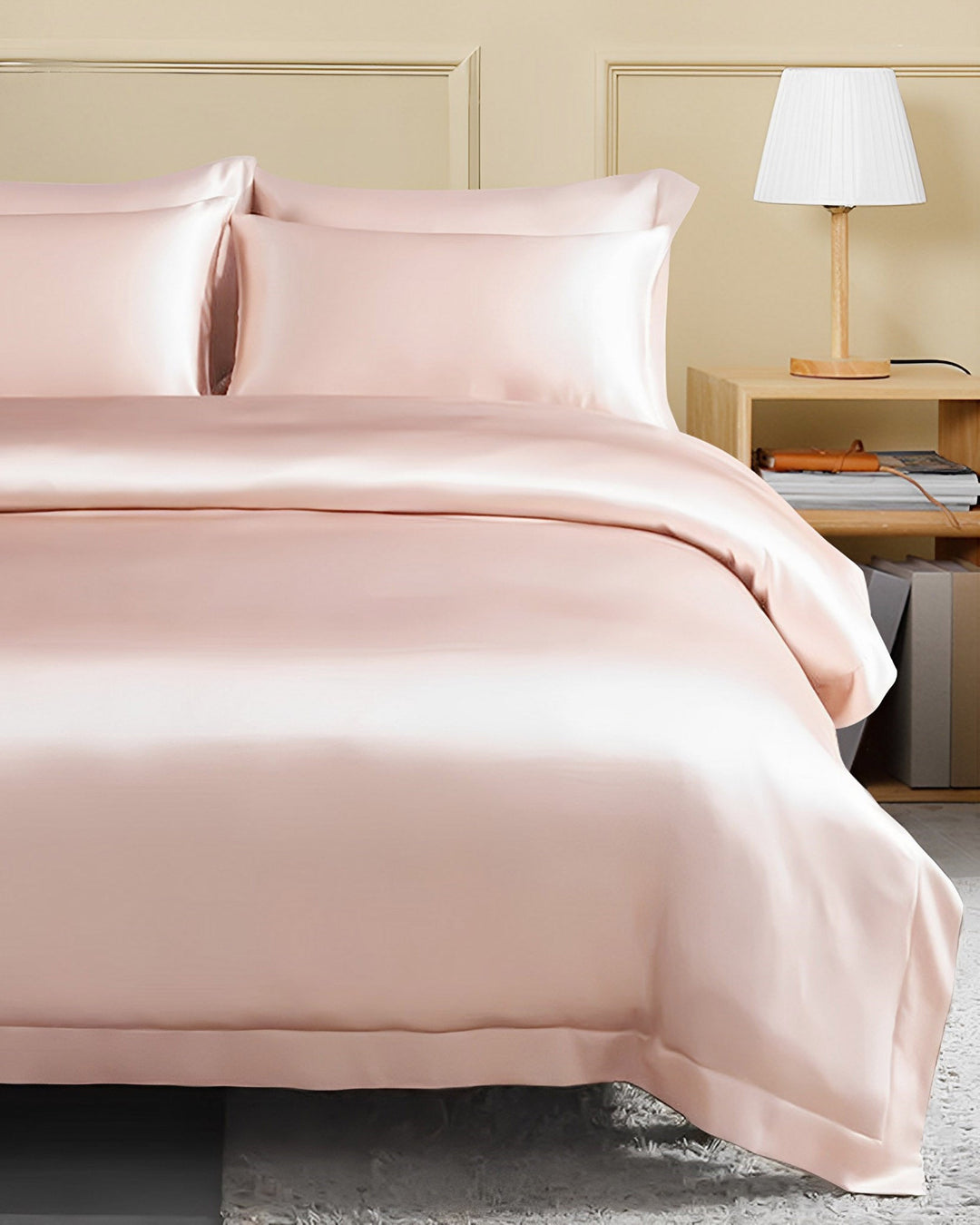 A neatly made bed features the 25 Momme Seamless Luxury Duvet Cover with Flange Border from SusanSilk, in a pale pink shade paired with matching pillowcases. Beside the bed, there is a wood side table adorned with open books and a white lamp. The wall behind the bed is decorated with elegant moldings.