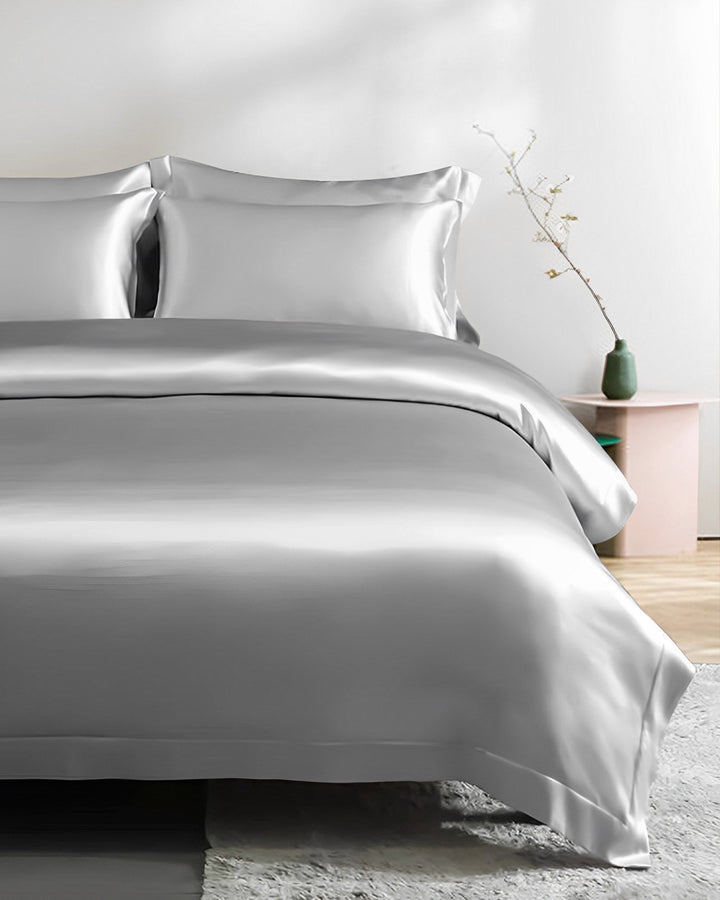 A neatly made bed features a 25 Momme Seamless Luxury Duvet Cover with Flange Border in light gray silk by SusanSilk and is adorned with two pillows. A small table holding a green vase with a thin branch stands next to the bed, completing the room's light, minimalist decor.