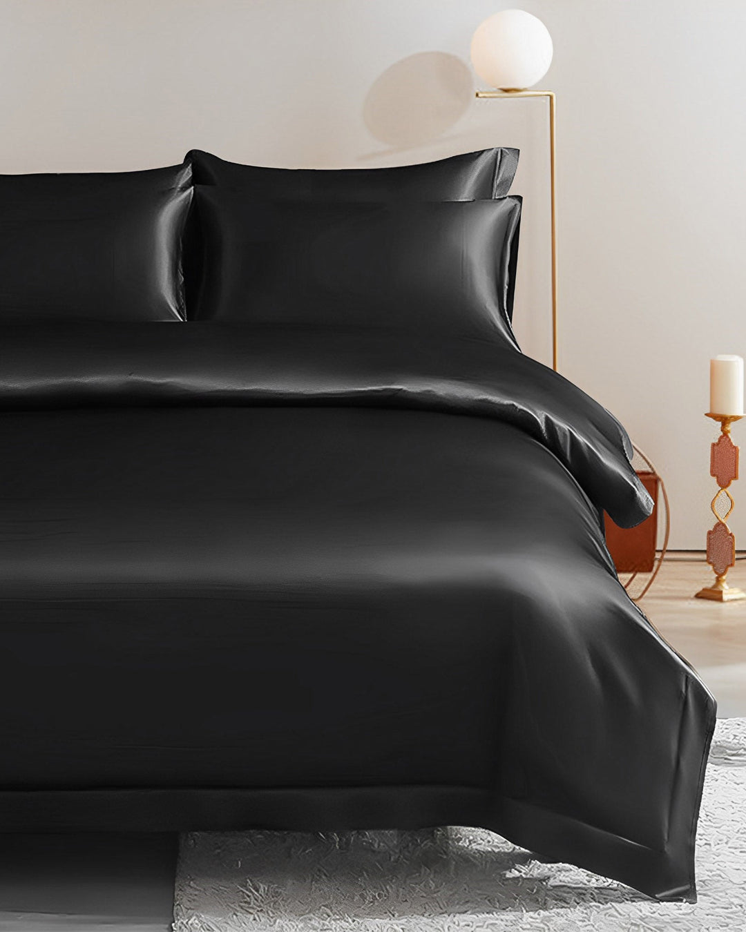 A neatly made bed with SusanSilk's 25 Momme Seamless Luxury Duvet Cover - Flange Border in black silk, including matching pillows and duvet. The room features minimalist decor with warm lighting, a modern lamp on the nightstand, and a candle stand in the background.