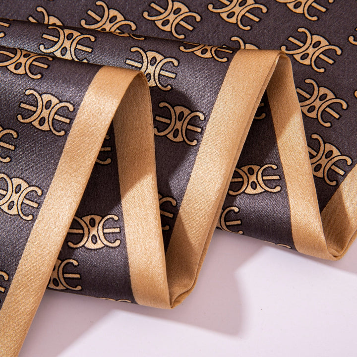 A neatly folded Arc De Triomphe Narrow Long Silk Scarf by SusanSilk, featuring a dark gray fabric adorned with a golden pattern and border.