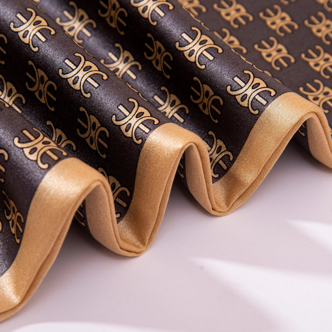 A close-up of the folded Arc De Triomphe Narrow Long Silk Scarf by SusanSilk, showcasing an intricate gold and black pattern with stylized symbols.