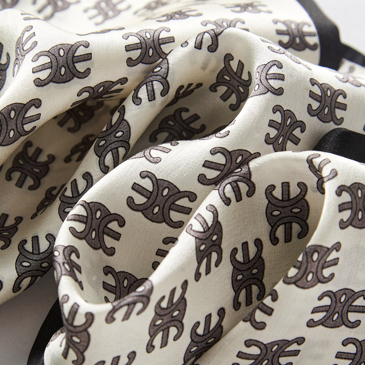 Close-up of SusanSilk's Arc De Triomphe Narrow Long Silk Scarf, featuring a cream-colored base adorned with a repeated black abstract symbol pattern. The luxurious silk fabric is gathered, showcasing its elegant folds and creases.