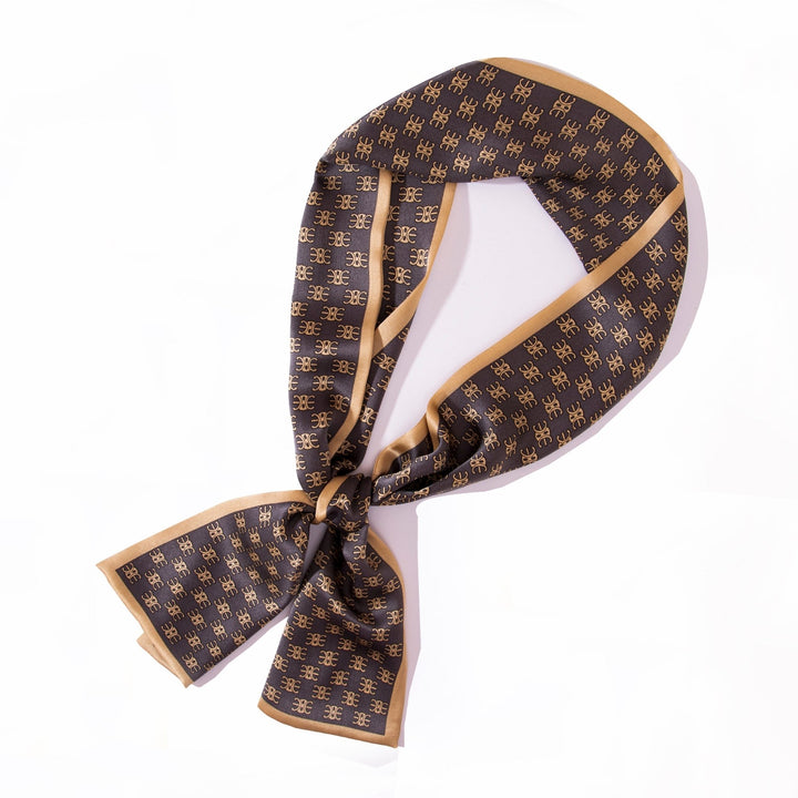 An elegant Arc De Triomphe Narrow Long Silk Scarf by SusanSilk, featuring a luxurious black and gold pattern, is tied in a loose knot and laid out on a pristine white background.