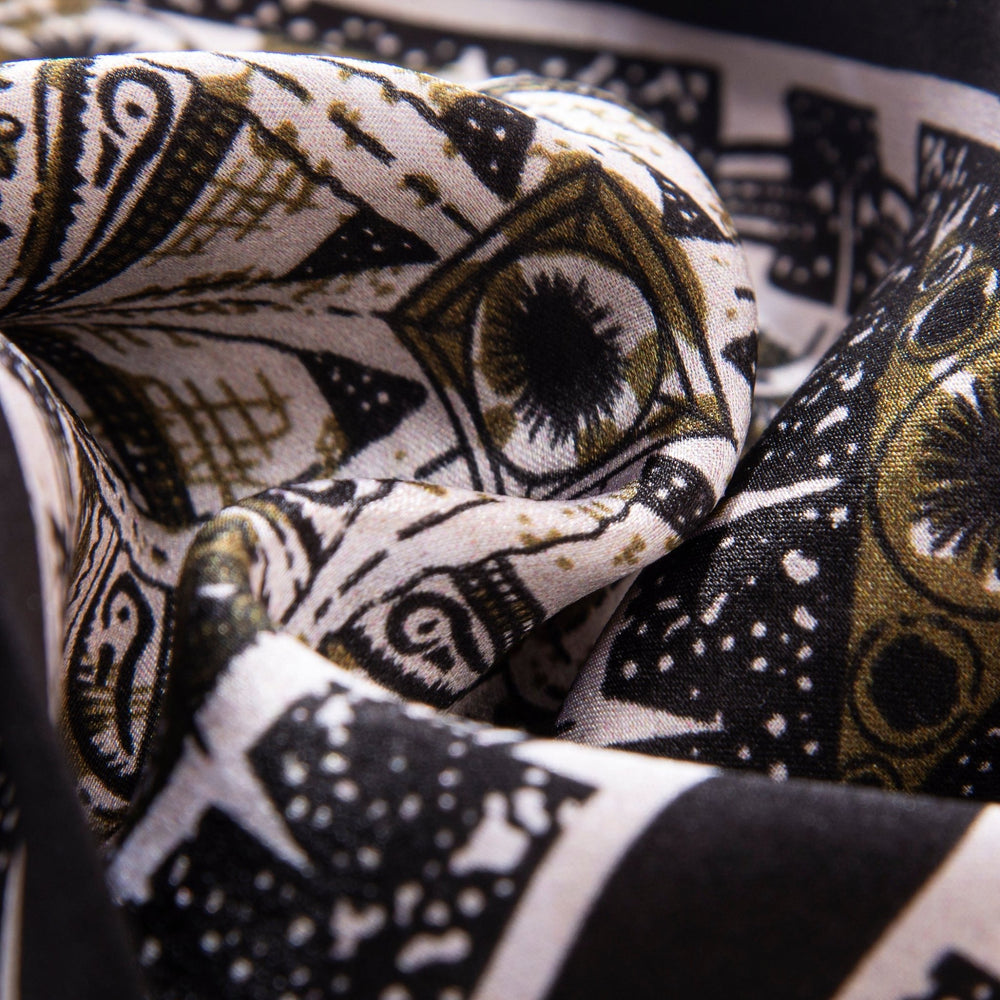 Close-up of a SusanSilk Black Compass Narrow Long Silk Scarf showcasing an intricate black and gold geometric pattern with eye designs and abstract shapes.