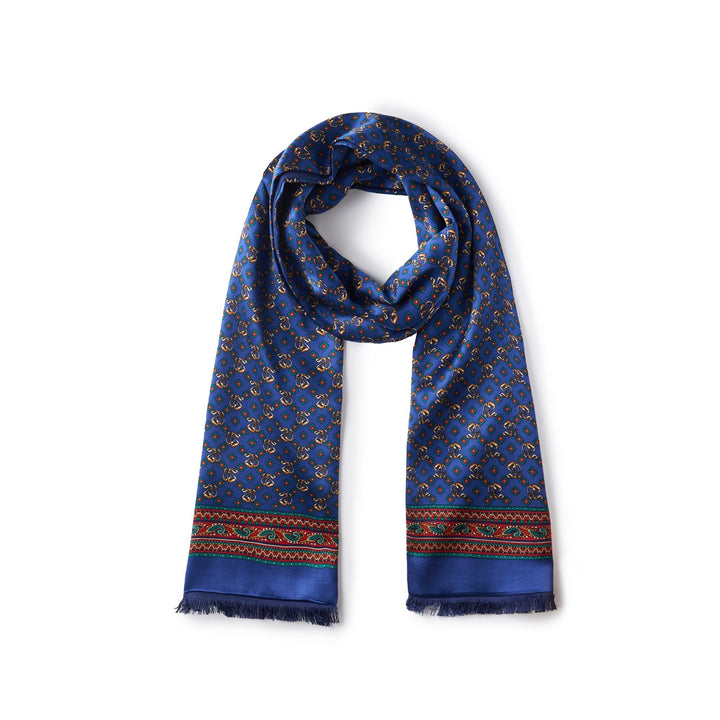 The Buckle Pattern Long Silk Brushed Scarf by SusanSilk is a long blue silk scarf featuring a repeating circular pattern and adorned with a decorative red, green, and yellow border near the edges.