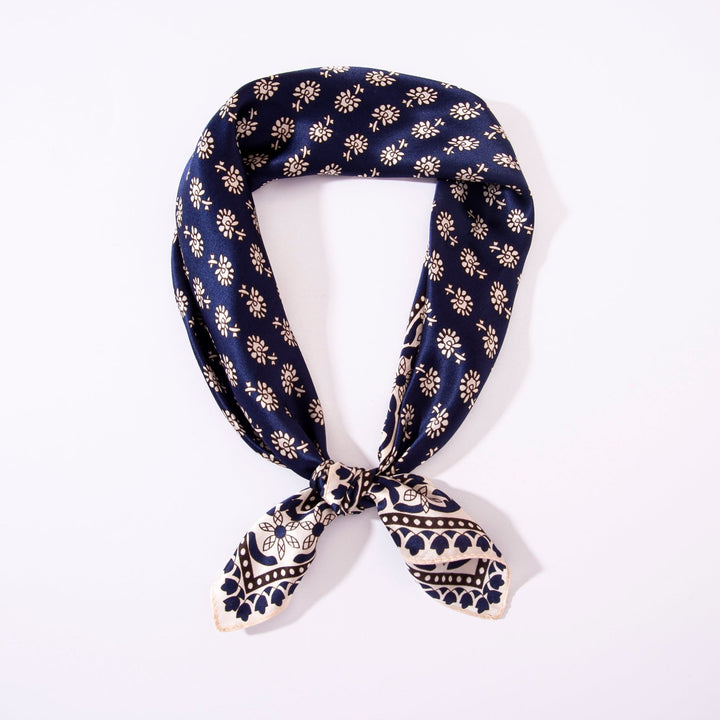 A blue Floral Pattern Square Silk Scarf from SusanSilk, featuring white floral and geometric designs, folded into a triangular shape and tied at the ends.