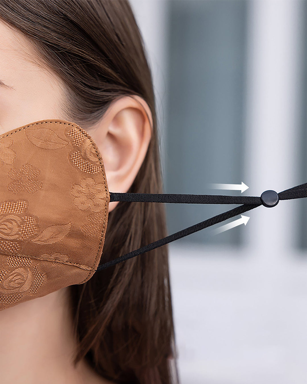 Close-up of a person wearing the SusanSilk Jacquard Upturned Brim Hat & Face Mask, featuring a luxurious brown patterned silk mask with adjustable black elastic straps highlighted by white arrows. The background is blurred.