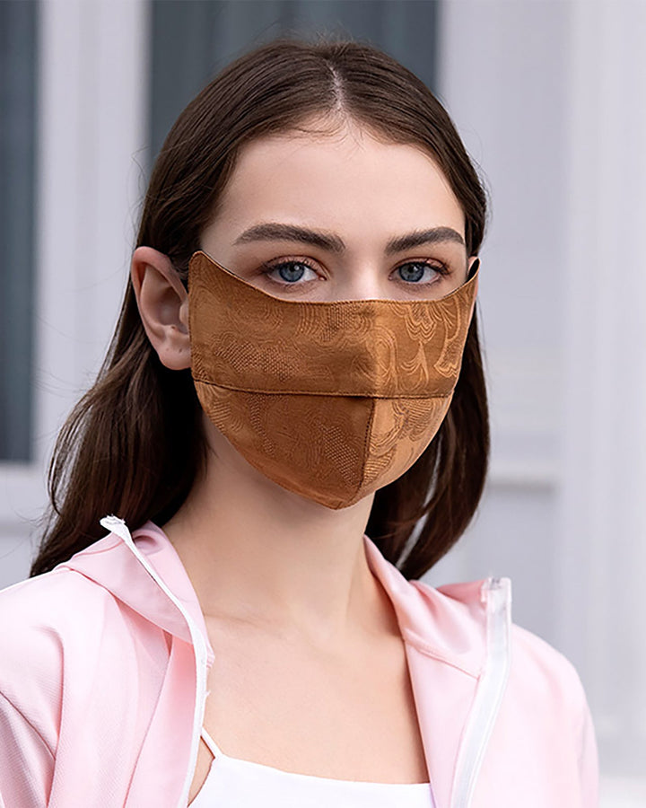 A woman with brown hair, wearing a Jacquard Upturned Brim Hat & Face Mask from SusanSilk in copper-colored silk, and a light pink jacket, looking directly at the camera.