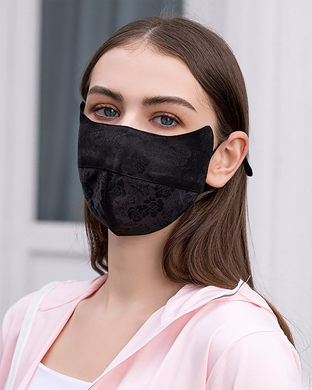 A person with long hair is wearing a black SusanSilk face mask from the Jacquard Upturned Brim Hat & Face Mask collection, paired with a light pink silk jacket, standing indoors with a neutral expression.