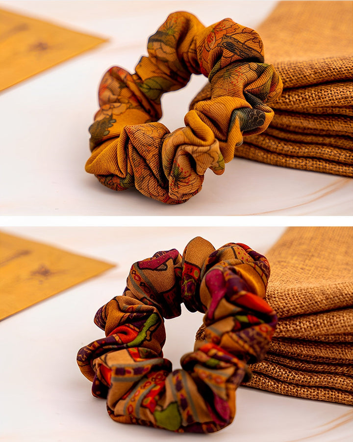 Two Gambiered Canton Gauze Silk Scrunchies from SusanSilk on a white surface. The top scrunchie is light brown with a subtle pattern, while the bottom one is a darker brown with an intricate, colorful design. Both are next to a piece of SusanSilk fabric, adding an extra touch of elegance to their appearance.