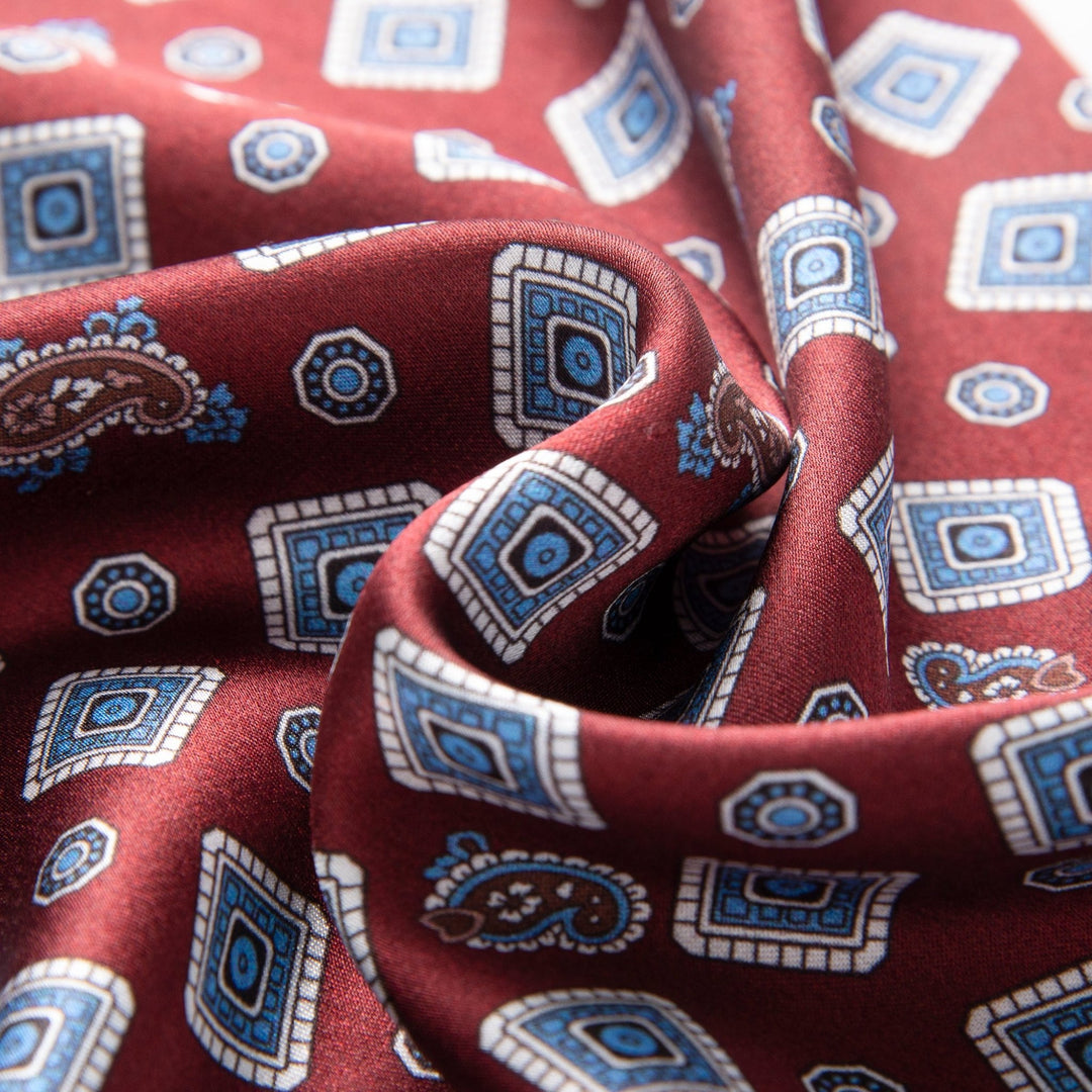 A close-up photo of the SusanSilk Gemstone Long Silk Scarf in burgundy, showcasing a pattern of blue and white geometric shapes alongside paisley designs. The luxurious fabric appears slightly wrinkled and folded.