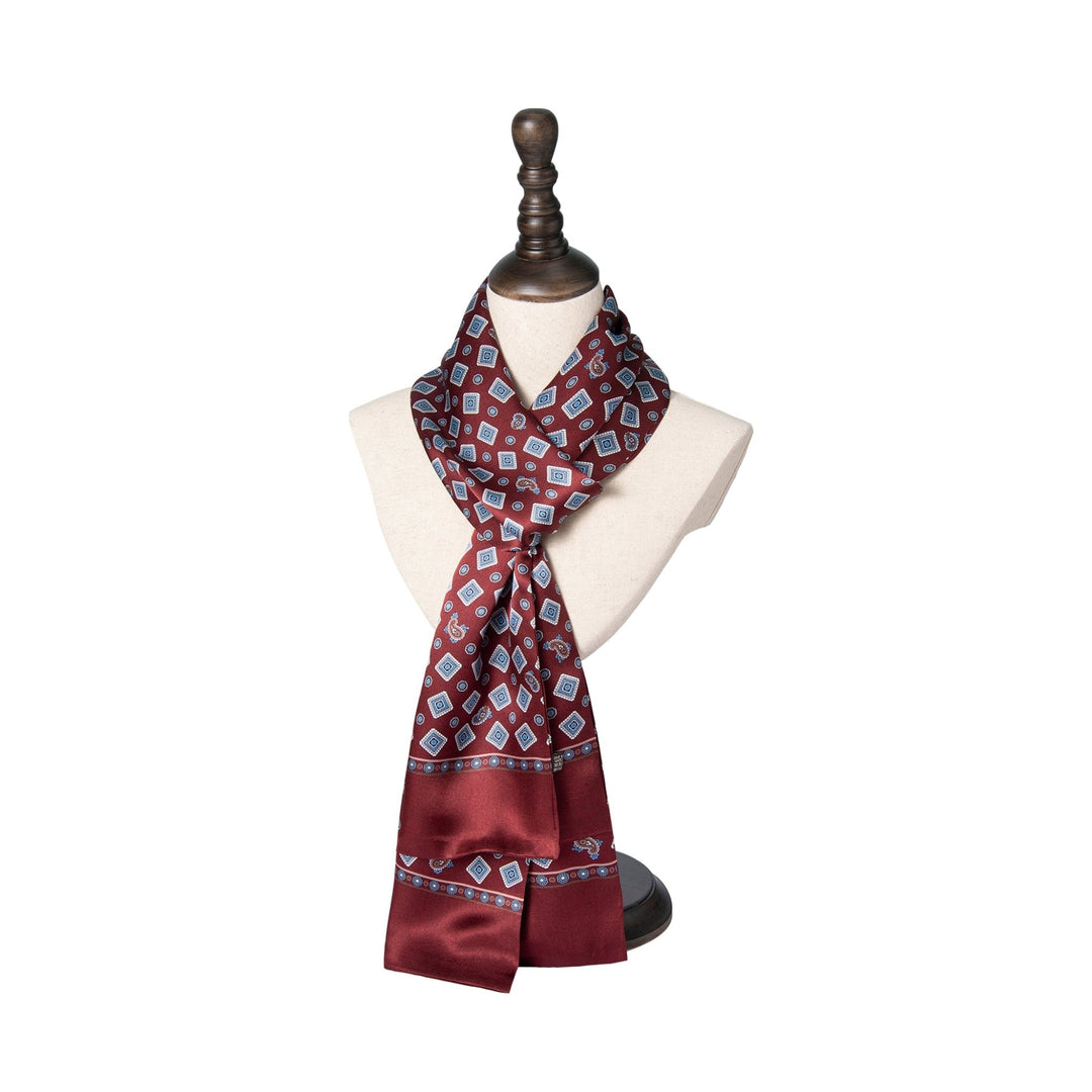 A maroon Gemstone Long Silk Scarf by SusanSilk, adorned with blue diamond patterns, is elegantly displayed on a white mannequin bust with a wooden stand.