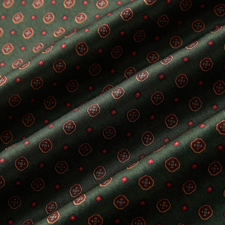 Close-up of the SusanSilk Geometric Pattern Long Silk Brushed Scarf in green. The dark green fabric showcases a pattern of small, evenly spaced decorative red and gold circles and squares. The luxurious silk material is slightly folded, creating gentle shadows and highlights.