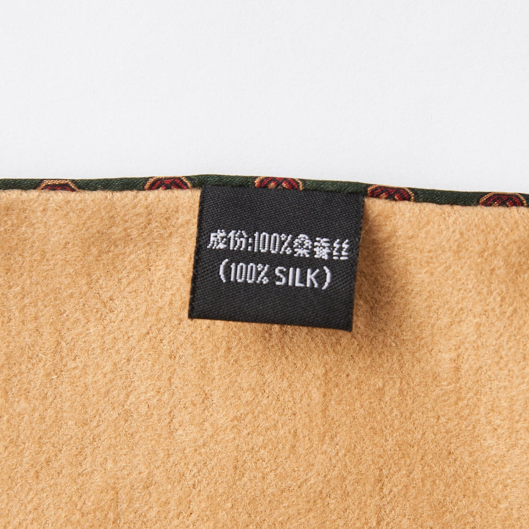 The SusanSilk label, displaying "100% Silk" in both English and another language, is attached to the Geometric Pattern Long Silk Brushed Scarf - Green, characterized by its tan-colored fabric with a striking green and red patterned edge.