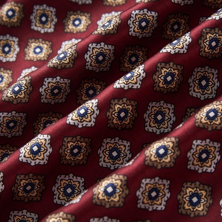 Close-up of a luxurious SusanSilk Geometric Pattern Long Silk Brushed Scarf in red, featuring a repeating pattern of small, ornate square designs in blue, yellow, and white.