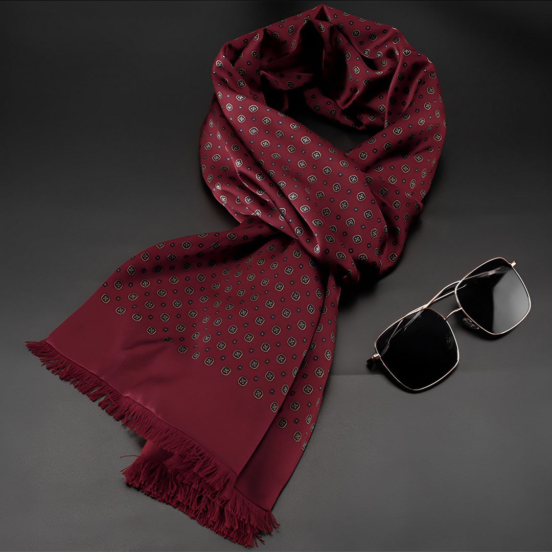 A Geometric Series Long Silk Brushed Scarf by SusanSilk, featuring a maroon base with white polka dots and fringed ends, is elegantly placed next to a pair of black aviator sunglasses on a dark surface.