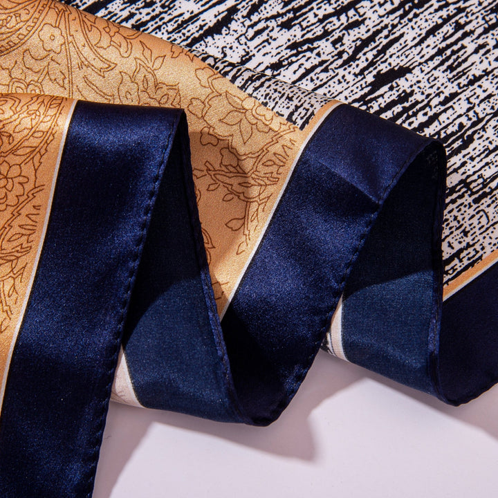 Close-up of a folded Jigsaw Series Square Silk Scarf by SusanSilk, showcasing a gold floral print with a dark blue satin border, elegantly displayed on a textured black and white background.