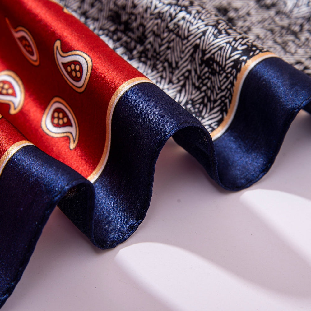 Close-up of the edge of a folded Jigsaw Series Square Silk Scarf by SusanSilk, showcasing its red and blue sections. The red part features a paisley pattern, while the blue edge has a wavy texture. The background is a white surface.